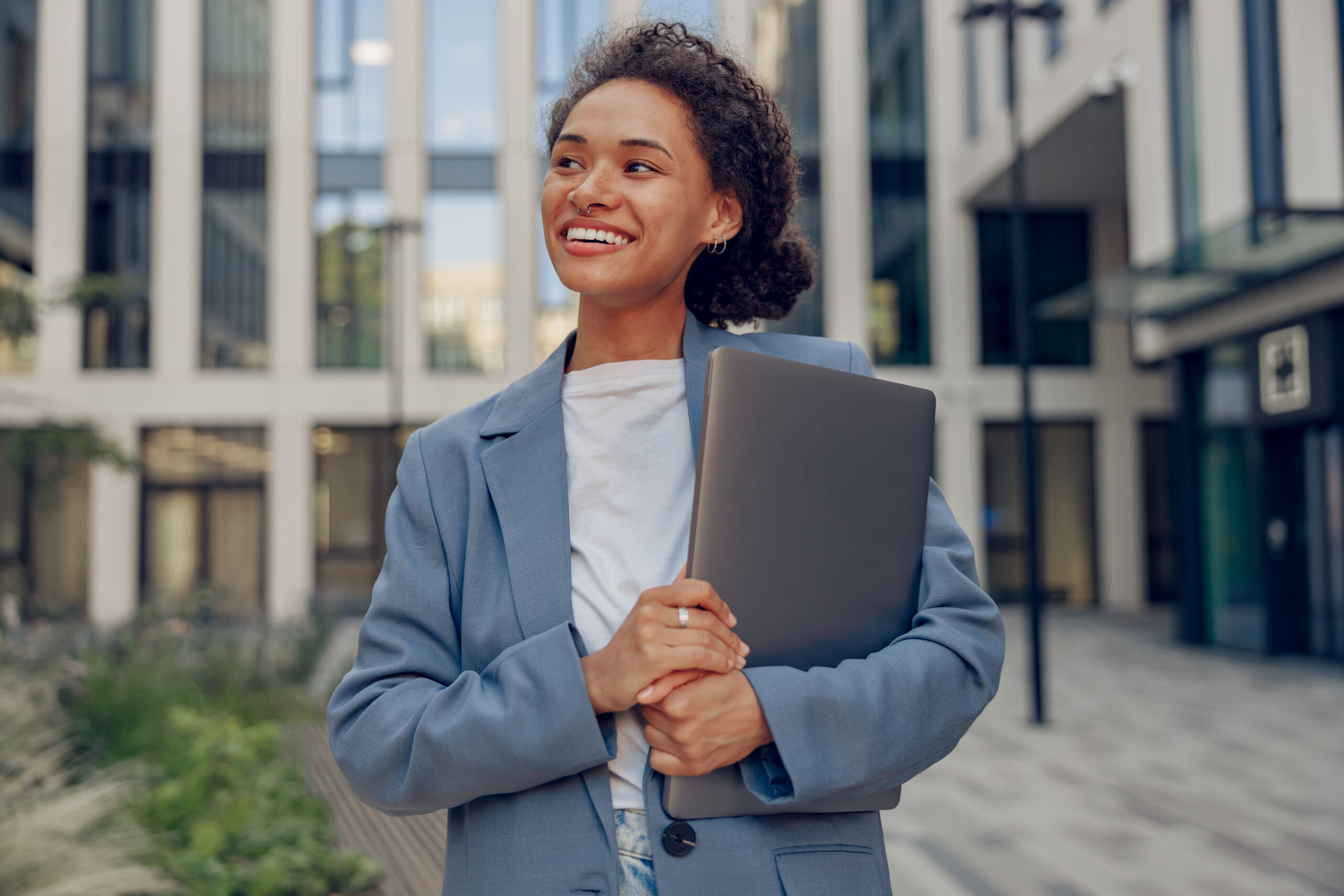 A smiling young professional holding a laptop while standing outside an office building